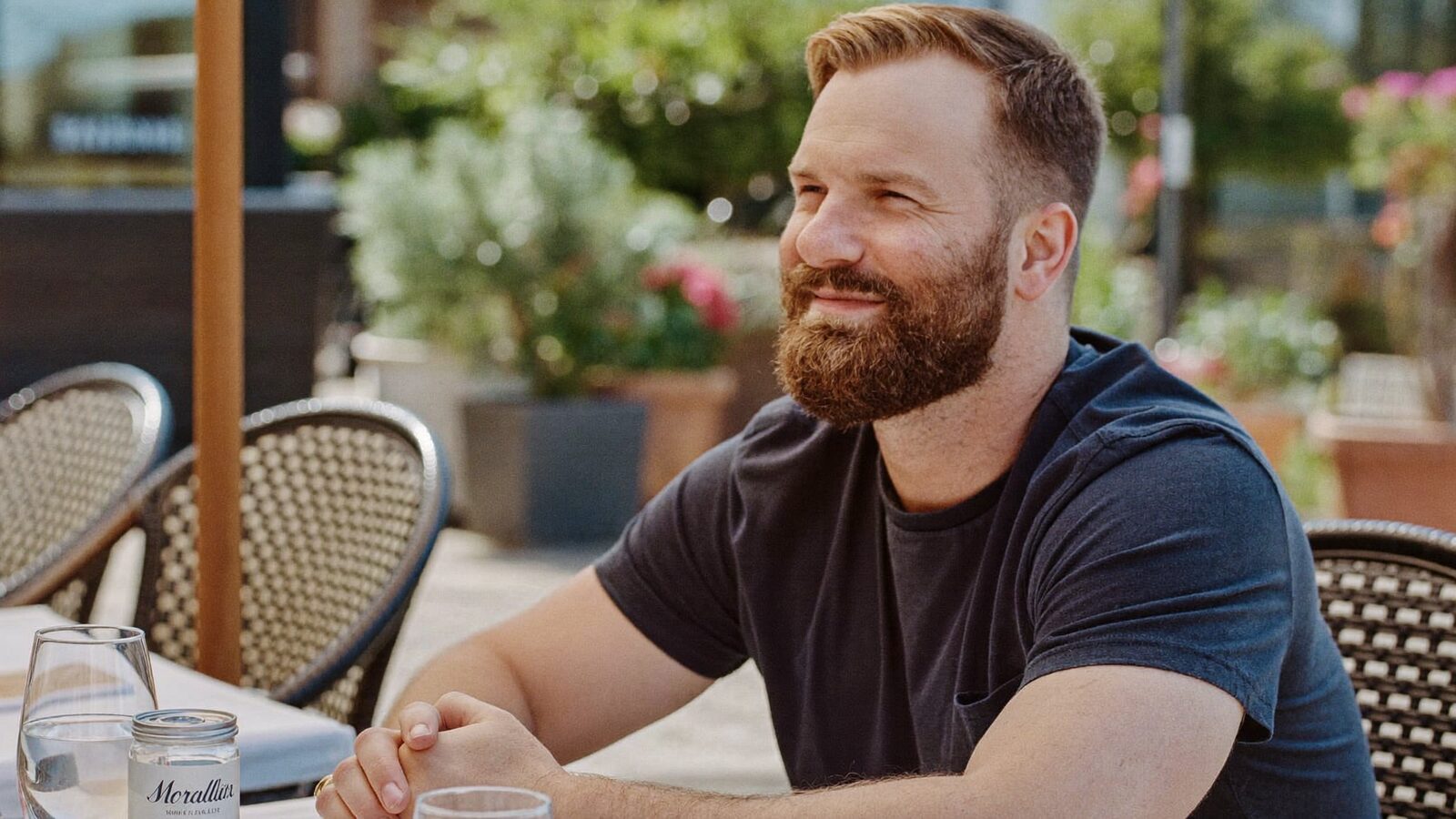 Man in his 30s with a dense, neatly shaped full beard sitting at a sunny outdoor cafe table