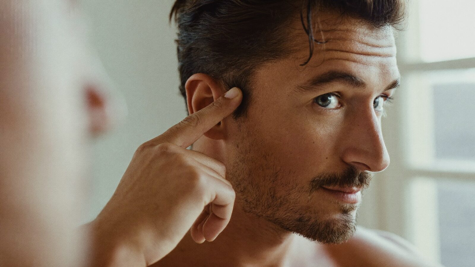 Man in his late 20s running a hand through his hair at the temple in a plain, bright room with soft window light