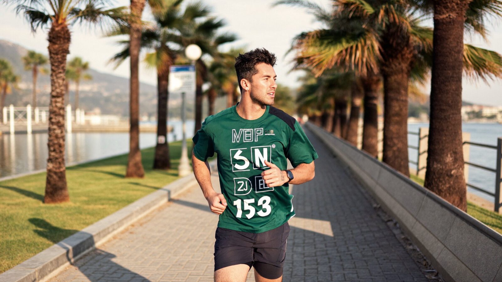 Fit man in his 30s with thick short hair jogging on a palm-lined waterfront path at sunrise