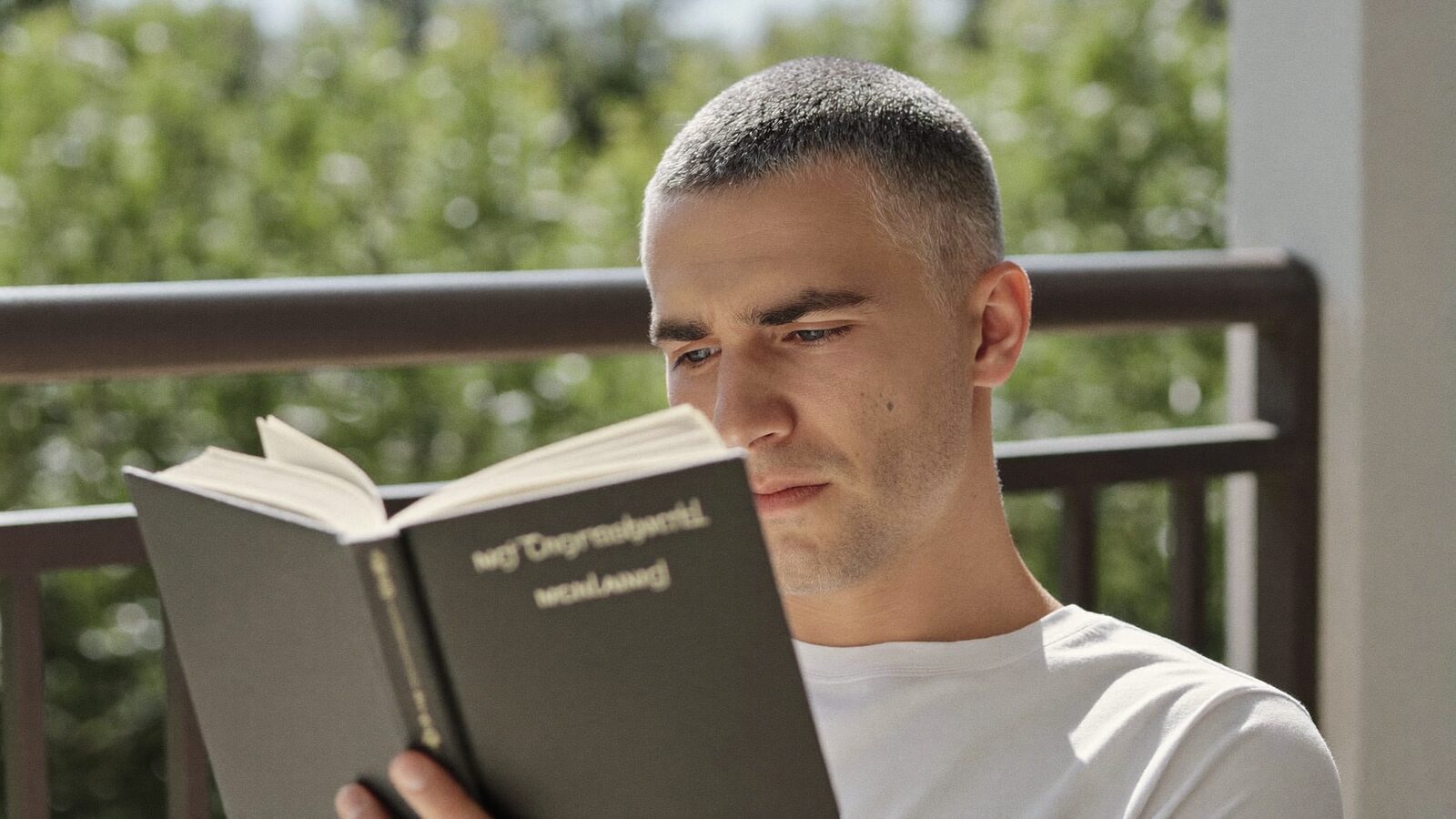 Man in his late 30s with a natural, full hairline reading a book on a bright balcony