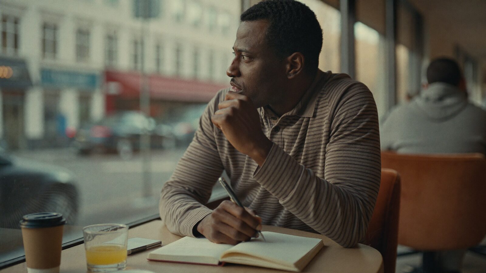 Man in his 30s with full natural hair thinking at a cafe window with a notebook in soft morning light