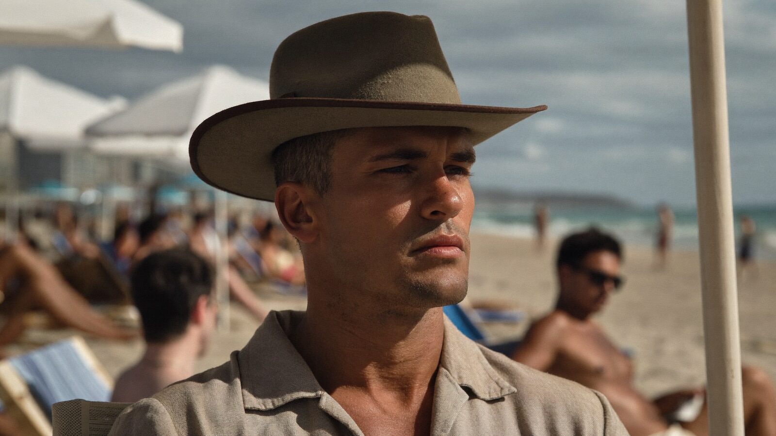 A man in his 30s with short healthy hair wearing a loose wide-brim hat, sitting in the shade of a beach umbrella in Miami