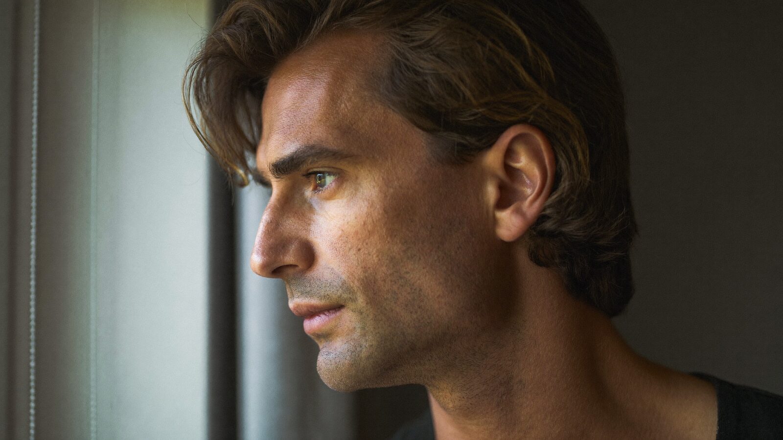 Man in his early 30s with slightly receding temples looking out a window, thoughtful, in soft morning light