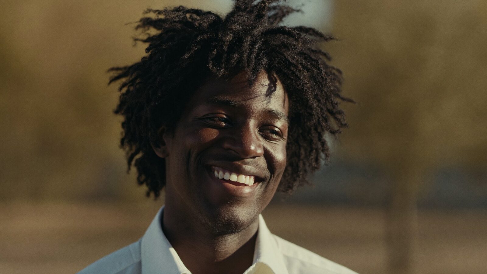 Black man in his 30s with a full, healthy curly hairline smiling outdoors in warm afternoon light