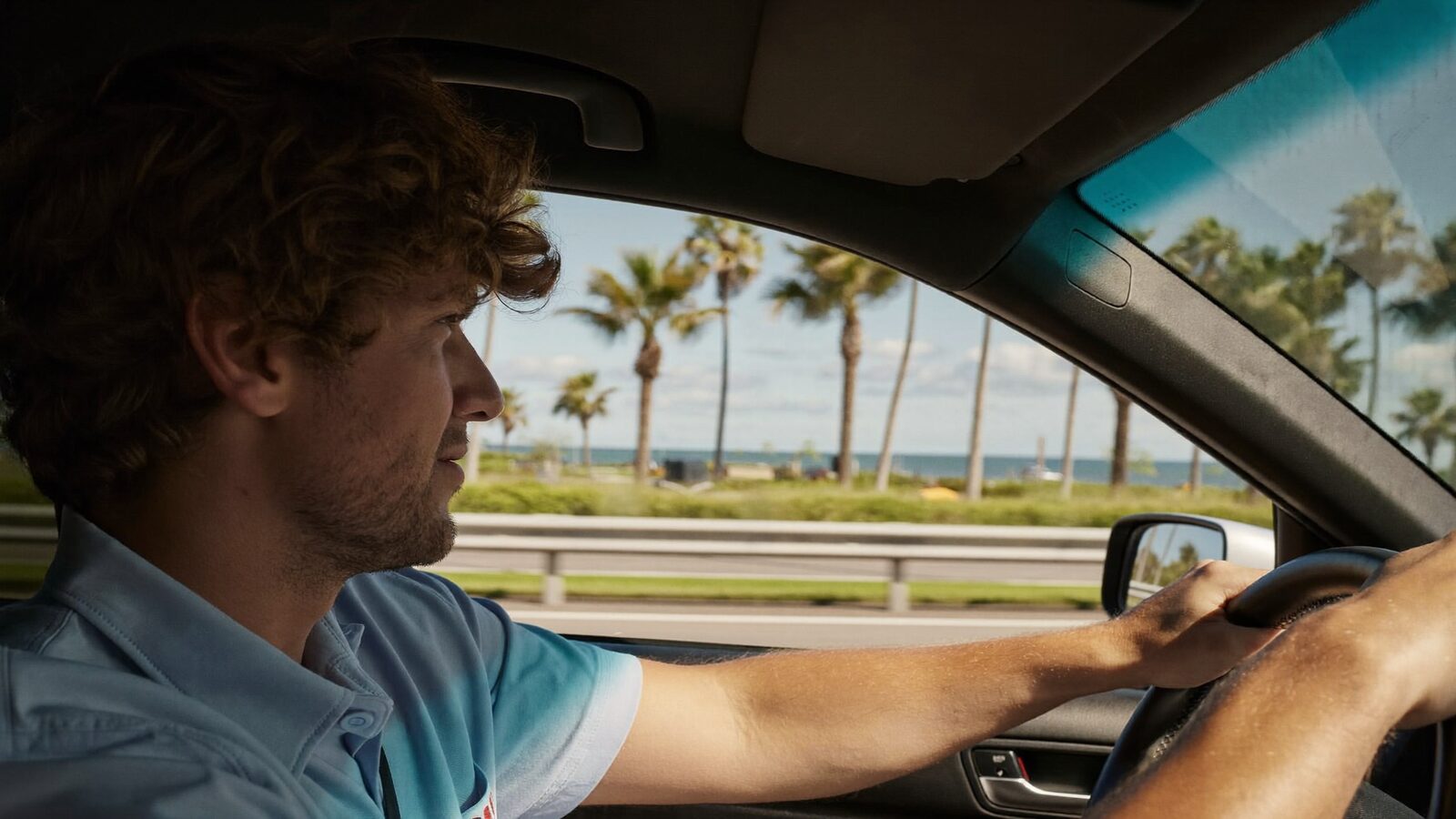 Man in his 30s with full hair driving a car along a sunny South Florida coastal highway, side view