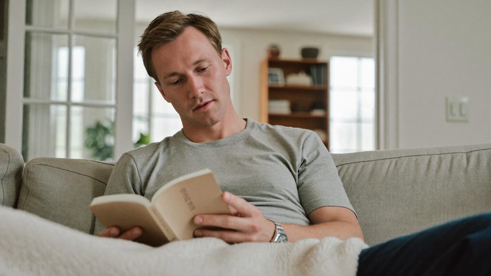 Man in his 30s with short neat hair resting on a sofa under a light blanket in a calm, bright living room