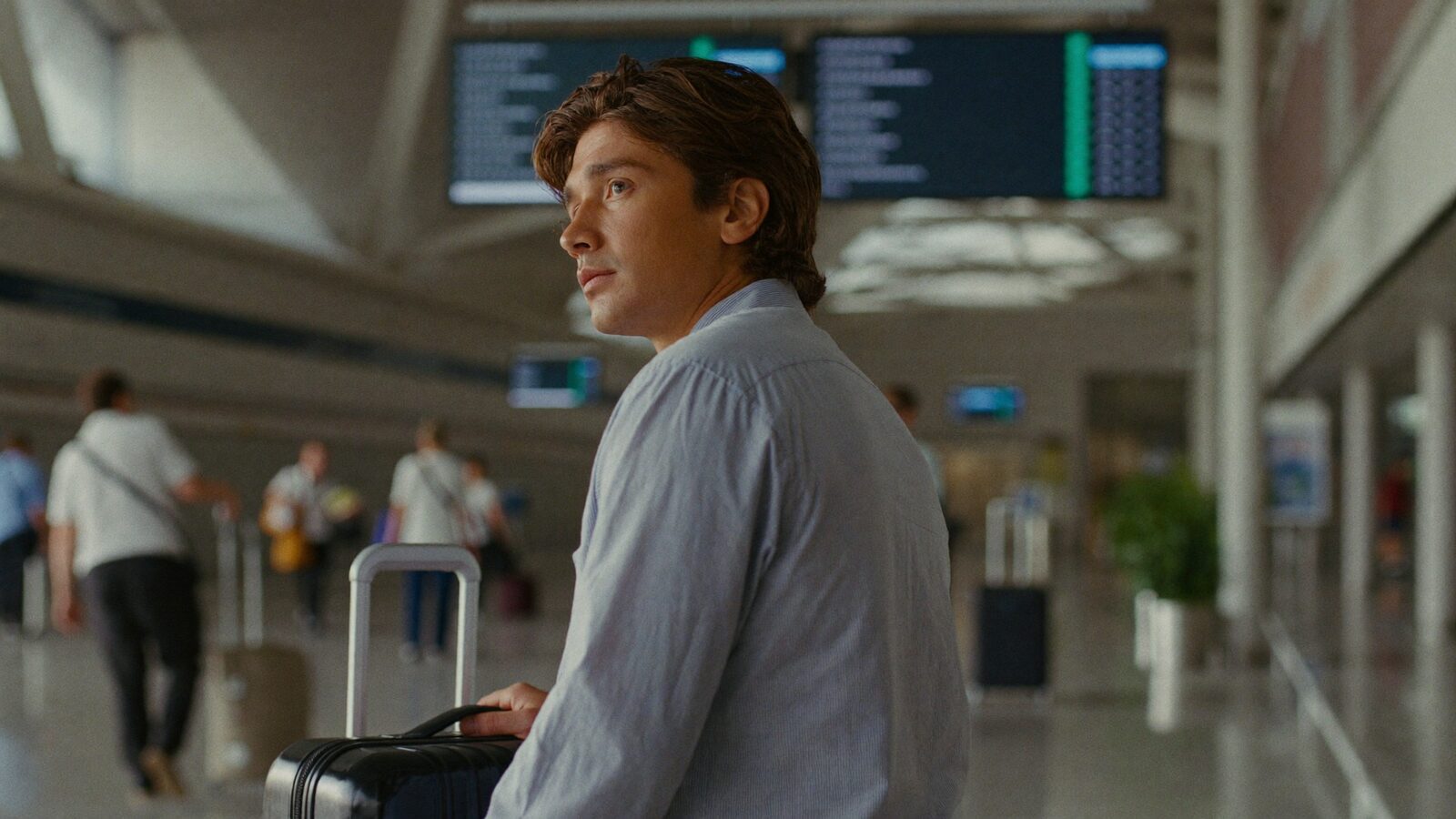 A man in his 30s with full hair looking at a departures board in an airport terminal, suitcase beside him