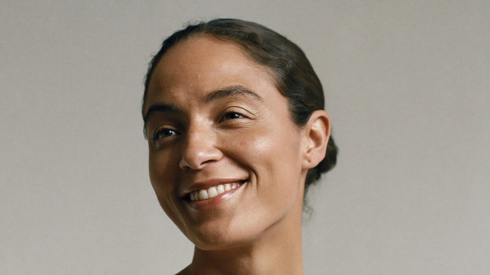 Woman in her 30s with a softly rounded natural hairline, hair pulled back, smiling against a plain light background