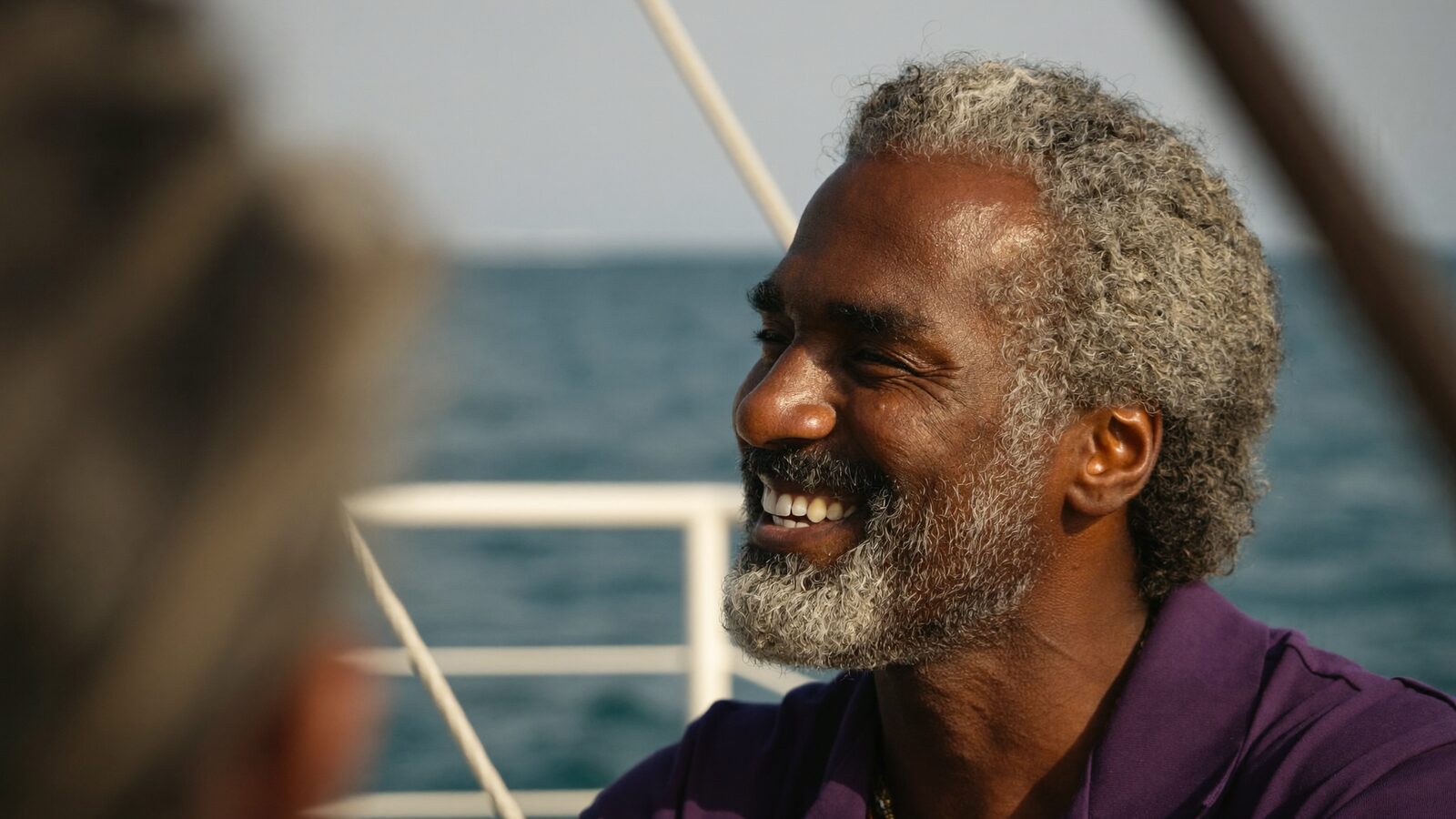 Man in his 50s with a full, natural salt-and-pepper hairline smiling on a boat deck with the sea in the background