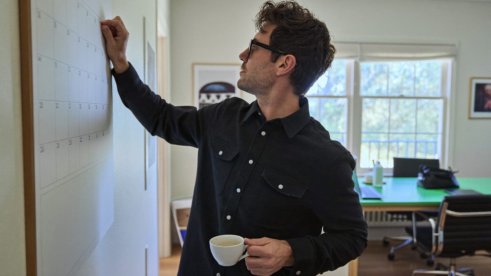 Man in his 40s with full hair checking a wall calendar in a bright home office, coffee cup in hand
