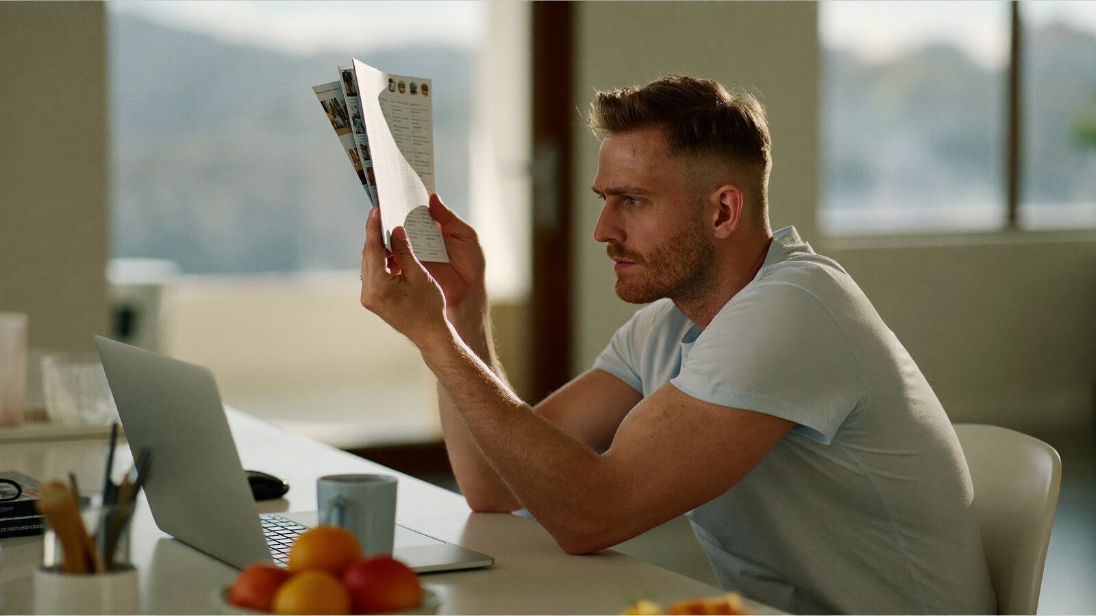 A man in his 30s with a thinning crown comparing printed brochures at a kitchen table with a focused expression