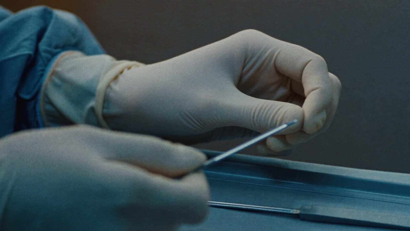 Close-up of gloved hands holding a fine handheld surgical instrument over a sterile blue tray in clean clinical light