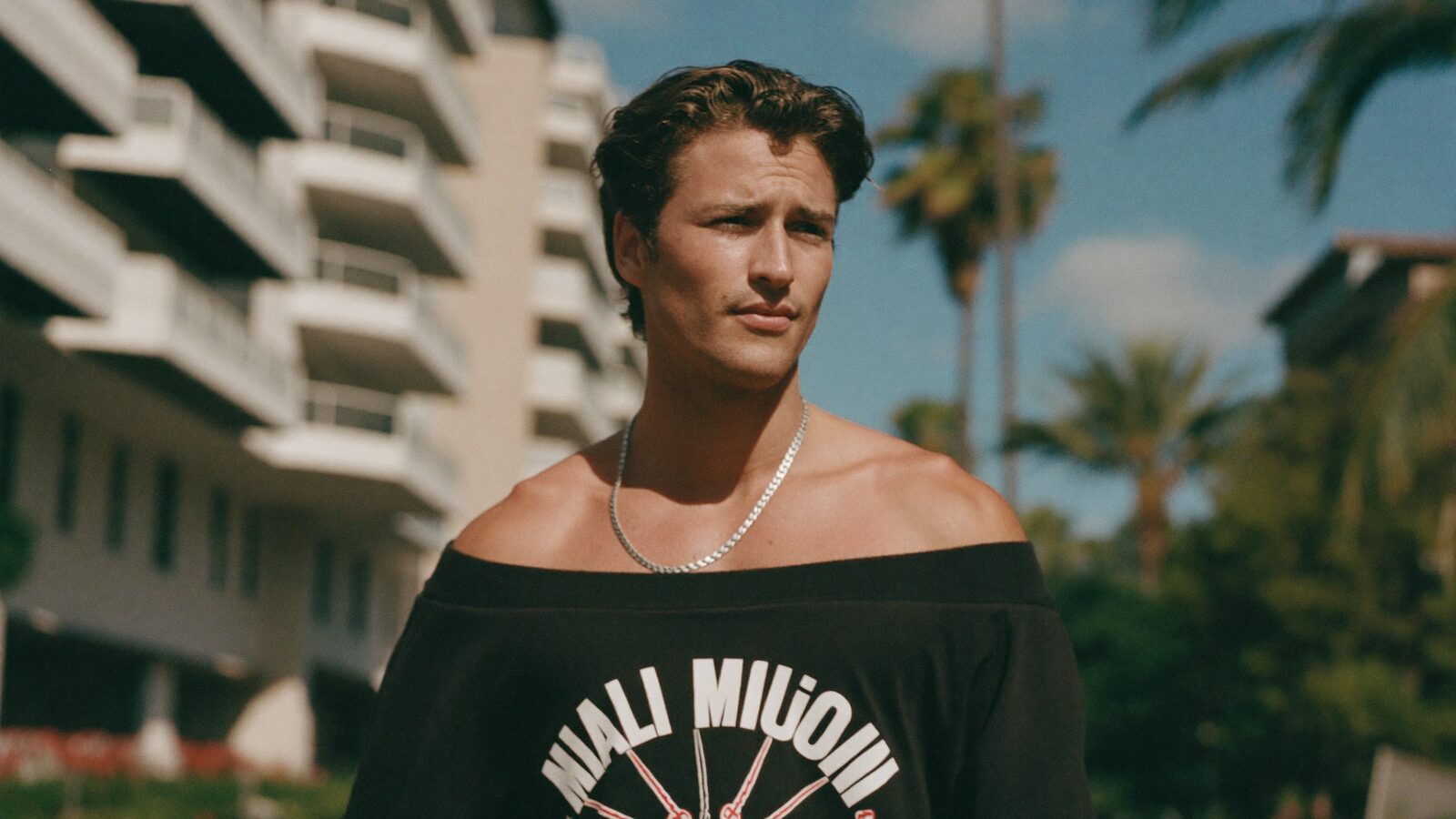 Man in his 30s with medium-length styled hair walking down a sunny Miami street, looking discreet and confident