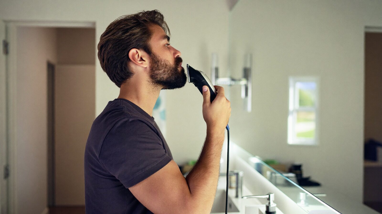 Man in his 30s trimming his full beard with clippers at a bright modern bathroom counter, side view