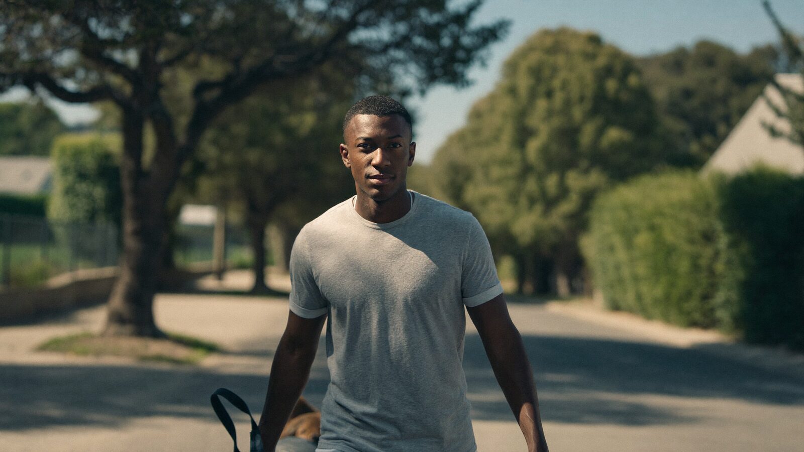 A man in his 30s with very short buzzed hair walking a dog along a quiet tree-lined street